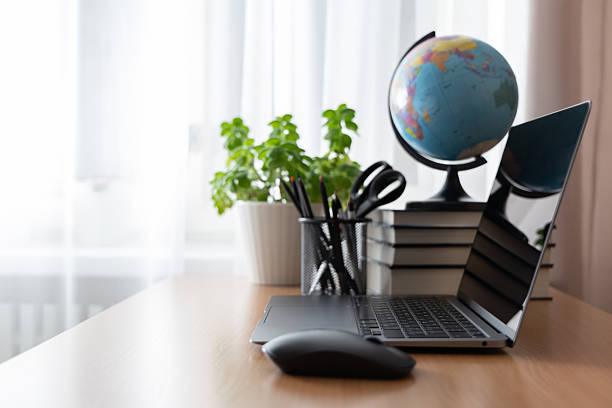 istock Stylish modern laptop on a desk, accompanied by a stack of books with a globe on top and a green potted plant nearby. Soft natural light from a window with white curtains creates a cozy atmosphere. 2249404789
