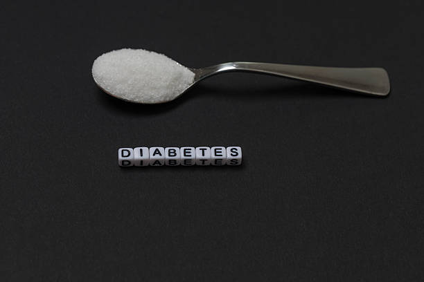 A chrome spoon full of white granulated sugar is centered above letter beads spelling out the word DIABETES. The image serves as a metaphor for the relationship between sugar consumption and the risk of diabetes