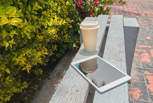 Coffee cup and digital tablet resting on a wooden park bench on a sunny day