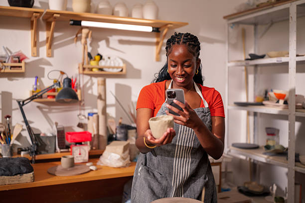 istock Young craftswoman smiling while taking pictures of a small clay pot in her workshop, using her smartphone to capture the perfect shot and looking at the screen with enthusiasm 2249419825