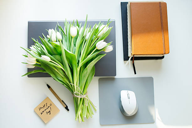 istock Sticky note with Women's day congratulations and white tulips bouquet laying at office workplace table with laptop, computer mouse, notepads. Sunny spring holiday morning greetings from colleagues. 2249421416