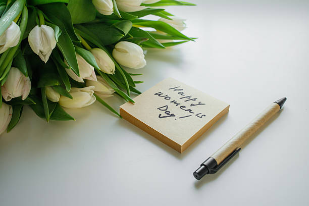 istock Sticky note with handwritten phrase happy women's day on white office table with pen and tulips flowers bouquet close up 2249421438