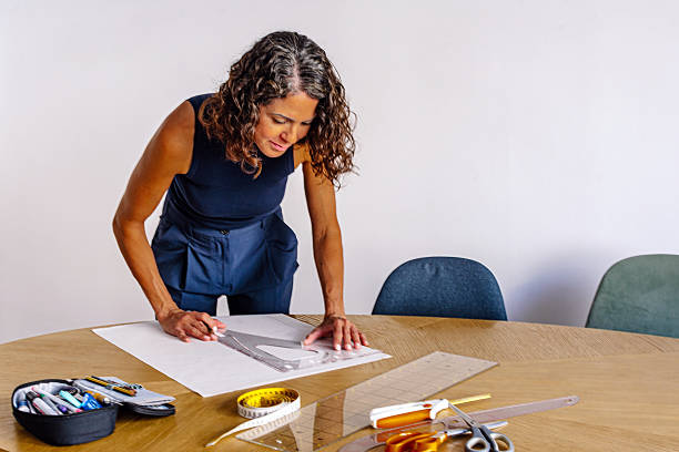 Woman concentrating on fashion design at a wooden table, sketching patterns and using ruler, tape measure, scissors and tools to craft clothing in a bright studio workspace