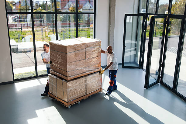 Two workers push a pallet of wrapped boxes through a bright, open office with large glass windows, demonstrating teamwork during an office move and logistics in a new workspace.