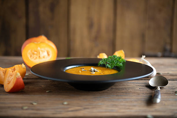 Warm pumpkin soup served in a black bowl with fresh parsley, with sliced pumpkin in soft-focus background. Cozy, autumn cooking atmosphere.
