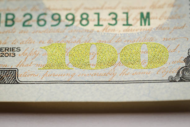 Close-up macro image of the yellow printed “100” denomination on a U.S. banknote, highlighting fine microtext, subtle color shifts, and textured paper fibers. The detailed view emphasizes the precision, security elements, and craftsmanship of modern currency design.
