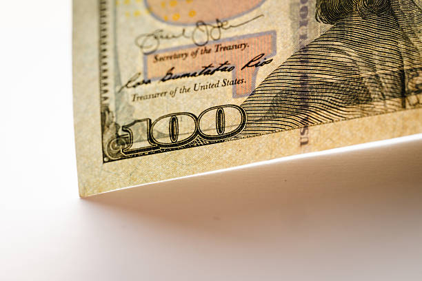 Close-up macro image of the lower corner of a U.S. hundred-dollar banknote, highlighting the bold “100” denomination, printed signatures, and fine engraving lines. The textured paper surface and subtle color elements emphasize authenticity and the craftsmanship of modern currency design.