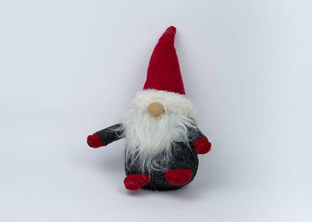 istock Plush gnome figure with a tall red hat sits against a white background. It has a white, fluffy beard, a round nose, and small red felt feet and hands. Christmas character. 2249449451