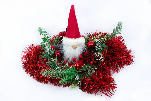 istock Christmas gnome decoration sits within an artificial green and red wreath. The plush gnome features tall red hat and white beard, surrounded by pine sprigs, baubles, and tinsel on a white background. 2249449481