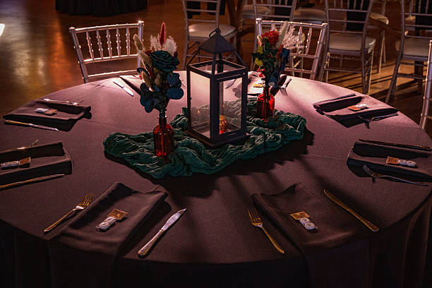 A close-up view of a wedding reception table decorated with dark linens, gold-toned silverware, and small floral arrangements in red glass vases. A lantern centerpiece sits atop textured fabric, with softly lit chairs and tables visible in the warm, rustic background.