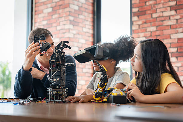 istock Mentor Guiding Students in Robotics and Virtual Reality (VR) Class, Man Showing Robotic Component to Girls with VR Headset, Children Learning Engineering and Programming with Adult Supervision 2249465316