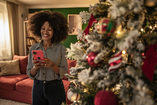istock Young black woman using phone next to Christmas tree at home on Christmas 2249479464