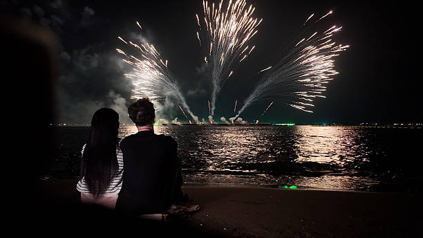 istock Romantic couple sits in a warm embrace, mesmerized by the dazzling fireworks display exploding over the sea, celebrating Christmas or New Year 2249483568