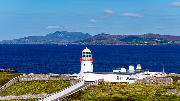 This image captures the remote and scenic St. John’s Point in County Donegal, located along the Wild Atlantic Way in northwestern Ireland. The view showcases the peninsula's lighthouse, an important maritime navigational aid built in the 1830s, surrounded by a stone enclosure and perched above rugged cliffs. The crystal-clear Atlantic waters and the jagged geology of the coastline emphasize the raw beauty of Ireland’s natural heritage. In the distance, the Derryveagh Mountains enhance the sense of remoteness and serenity. This landscape is ideal for travel-related visuals, nature documentaries, and editorial uses reflecting coastal life, exploration, and seafaring traditions.