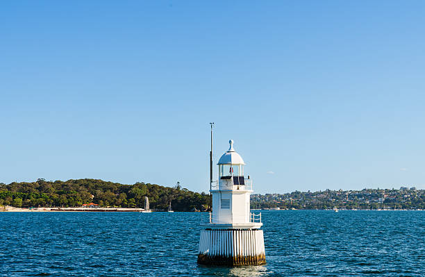 White lighthouse standing in the water with coastal buildings and forested shoreline in the distance on a clear sunny day in Sydney, Australia.