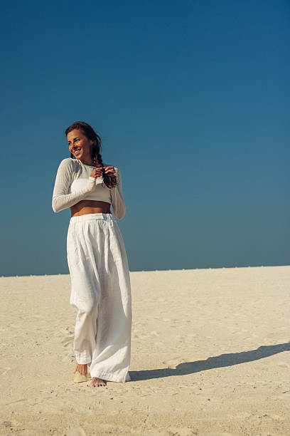 A carefree woman strolls barefoot on pale sand, wearing a white cropped top and wide-leg linen pants. The bright coastal scene features a deep blue sky and tranquil horizon, conveying relaxation, freedom, and summer travel. Perfect for lifestyle, wellness, and vacation themes.