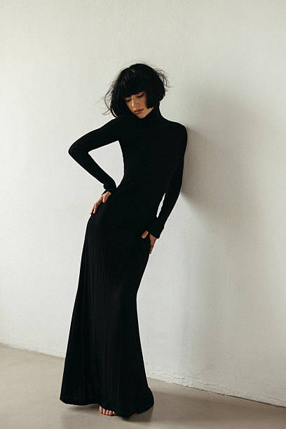 A fashion portrait featuring a woman in a sleek black turtleneck maxi dress, standing in a minimal studio against a white wall. The pose exudes elegance and confidence with a moody, serene vibe. Clean lines, monochrome palette, and soft lighting highlight the garment and silhouette.