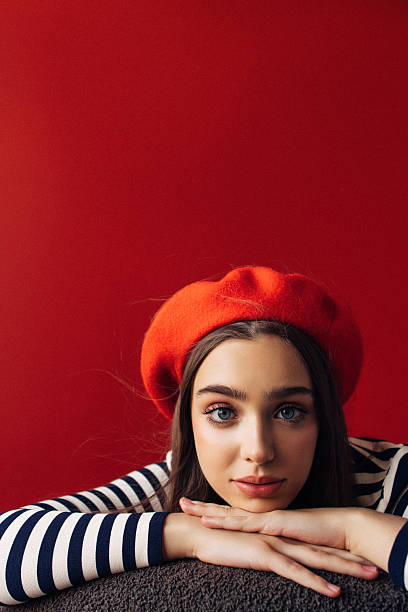 A bold, fashion-forward portrait featuring a young woman in a bright red beret and navy striped shirt. She rests her chin on her folded hands, creating a relaxed, playful mood. The vivid red background enhances the striking look, making it ideal for fashion, lifestyle, and creative branding projects.