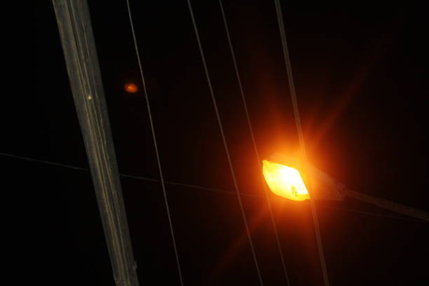A close-up view of a glowing orange street lamp at night, with dark electrical cables and a utility pole creating a stark, abstract pattern against the black sky.