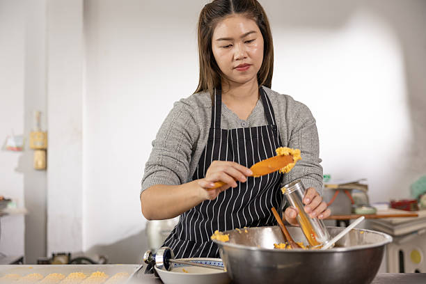 istock Asian Young entrepreneur in striped apron scooping dough into tin while standing at stainless steel bowl in bright kitchen preparing baked goods with focused expression and warm atmosphere 2249497448