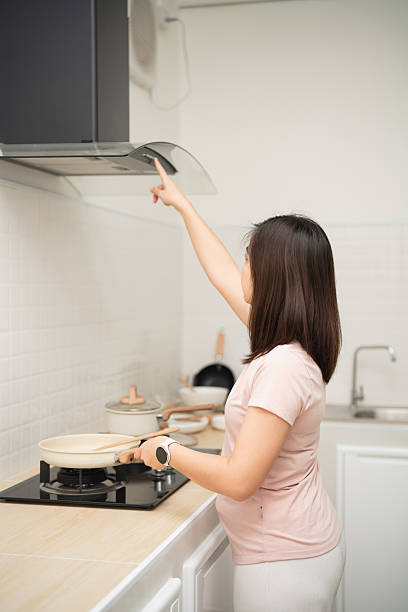 istock Young woman in a light pink shirt cooking in a modern kitchen, adjusting the range hood while preparing a meal on the stove with focus and care 2249498541