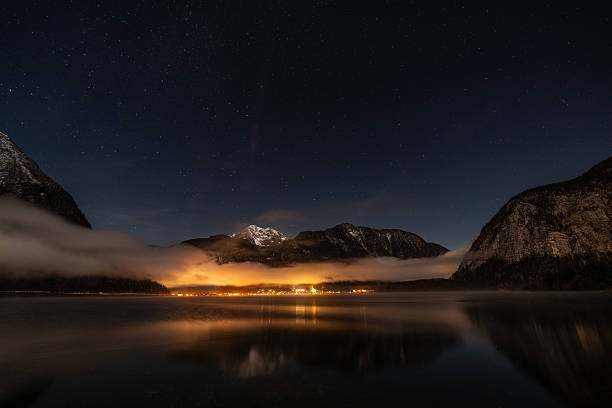 silent star clear night in austrian alps, calm lake Hallstatt, small village illuminated, some wafts of fog, a bit of snow on mountain peak