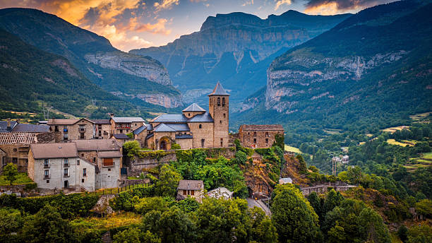 Aerial view of the historic village of Torla at sunrise in the Ordesa Valley of the Spanish Pyrenees. The scene features the iconic Church of San Salvador and surrounding stone buildings perched above lush green hills, with the towering Monte Perdido massif rising dramatically in the background beneath the glowing sunrise sky. Torla-Ordesa in Aragón, in the province of Huesca, northern Spain - Europe