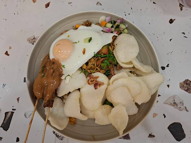 An appetizing overhead shot of a special Indonesian dish, featuring fried noodles/rice topped with a fried egg, satay in peanut sauce, and crispy prawn crackers.