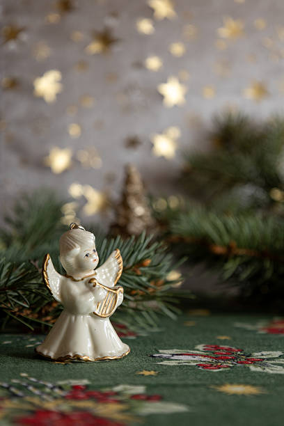 A delicate ceramic angel figurine holding a small harp is placed among fresh evergreen branches. Soft golden star-shaped bokeh lights create a warm festive backdrop. The scene conveys a peaceful and traditional Christmas atmosphere.