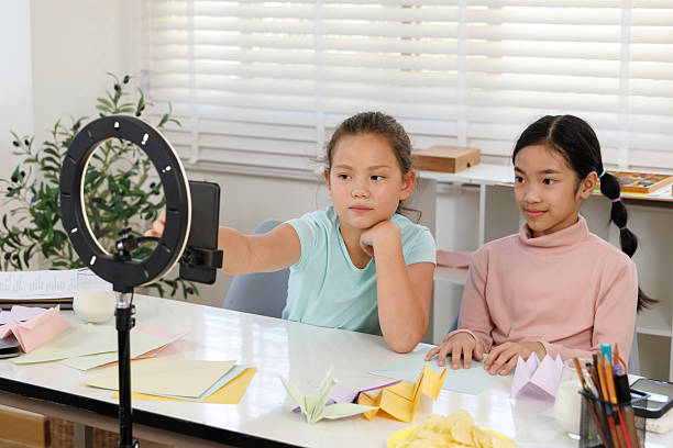 girls are using a smartphone and a ring light to film an origami tutorial video. Colored paper and finished paper crafts are on the desk, showcasing the creation of online content about handicrafts to share with viewers.