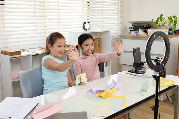 istock Two girls are live streaming or recording an origami tutorial video using a mobile phone and a ring light. There are colored papers and completed paper crafts on the desk. One girl is waving to the viewers, 2249506415