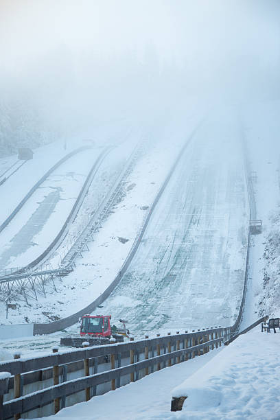 Snow removal vehicle cleaning ski jumping hill in winter fog. Maintenance work on sport infrastructure before ski jumping competition. Winter sports preparation, extreme sports facility, cold weather, alpine landscape and seasonal service concept