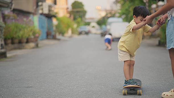 four-year-old Asian child learns to skateboard with guidance from his older sister during the early evening after school. The moment captures family bonding, childhood development, active outdoor play, and supportive sibling relationships. Perfect for themes related to parenting, education, sports learning, childhood lifestyle, evening activities, and family connection.