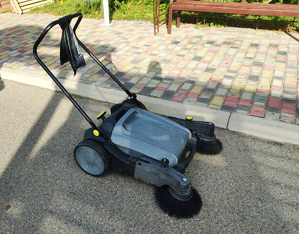 istock Manual push sweeper machine for outdoor floor cleaning, gray body and black handle, positioned on asphalt path near decorative paving stones and green garden area with wooden bench under daylight 2249514775