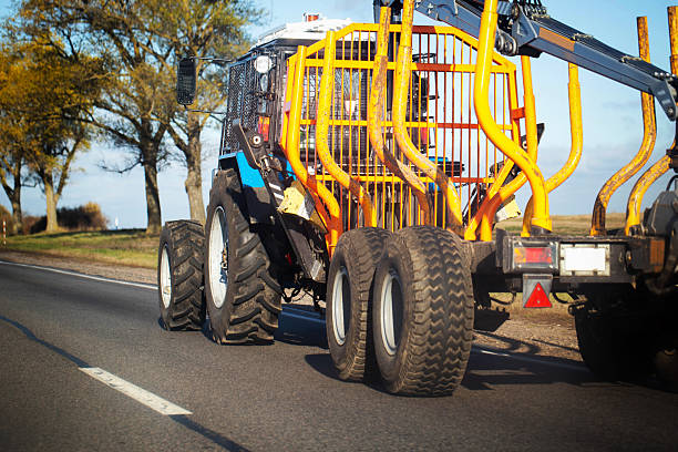 Rear view of powerful blue and yellow forest forwarder. Logging machine moves on rural asphalt road. Large rugged tires, robust safety cage, hydraulic arm for timber handling visible. Autumn trees line roadside, clear sky.