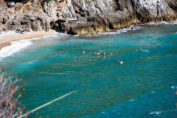 Varcaturo â Campania â Italy â 08-24-2025: Swimmers in turquoise coastal water near a rocky beach as small waves roll toward the shore and sunbathers rest on the narrow strip of sand.