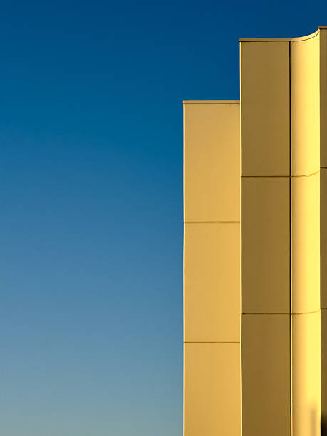 Sunlight casts warm hues and sharp shadows on a contemporary building's facade, featuring straight and curved lines under an azure sky.