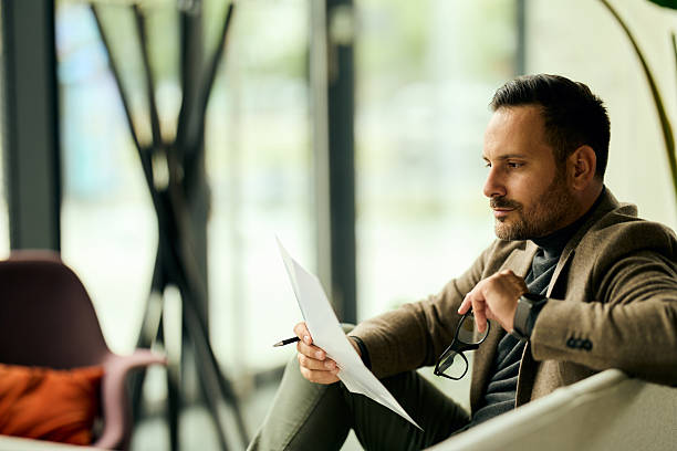 istock Professional Man In Blazer Reads Documents While Holding Glasses In Modern Office Lounge 2249531174