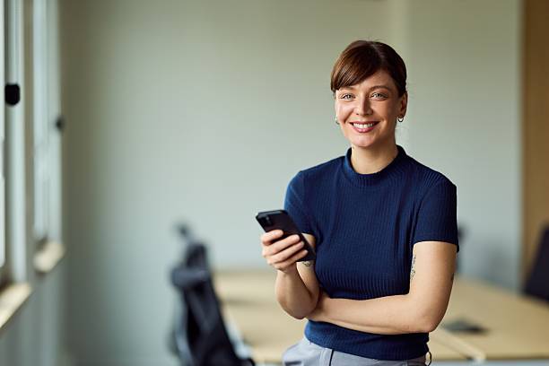 istock Confident Woman In Blue Knit Top Smiles While Using Smartphone In Modern Office 2249531185