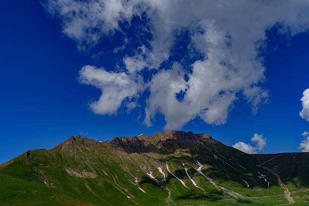 White clouds float above a jagged green ridge, casting shifting shadows across the mountain slopes.