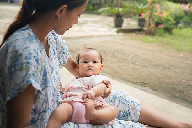 A baby sits on an adult’s lap and is spoon-fed in a quiet outdoor area with greenery