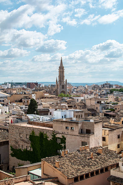 A bright panoramic view over the rooftops of Palma, Mallorca, showcasing warm-toned traditional buildings and the striking spire of a historic church rising at the center of the city. Soft clouds drift across a blue sky, adding depth and calm to the Mediterranean scene, while the mix of old stone architecture and modern structures highlights the city’s blend of heritage and contemporary life.