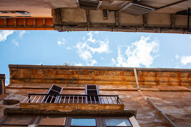A dramatic upward angle showing weathered stone façades, open shutters, and the bright sky above Palma’s historic old town. The warm textures and narrow gap between buildings capture authentic Mediterranean charm.