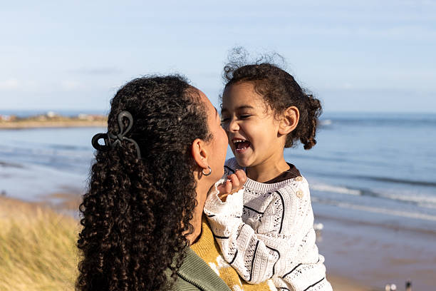 A waist-up shot of a mother carrying her young daughter along the coast on a beautiful, sunny day. They are both smiling, enjoying their time together by the ocean. Located in Northumberland, England.Videos are available of this scenario