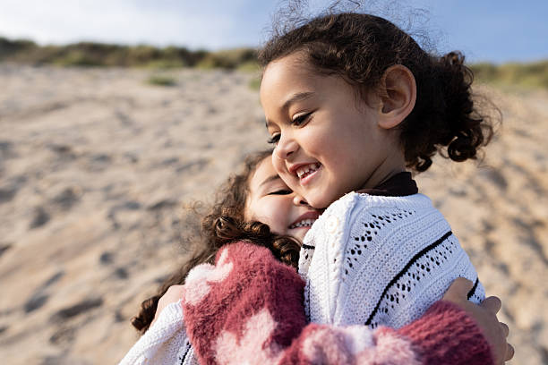 A close-up of two young sisters embracing lovingly on a sandy beach on a sunny day. They are both smiling, wearing casual clothing. A tall dune with a vibrant blue sky is in the background. Located in Northumberland, England.Videos are available of this scenario