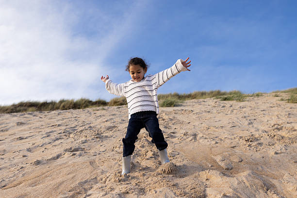 Girl with winter clothes running down a sand dune carrying a toy airplane, leaving footprints in the sand