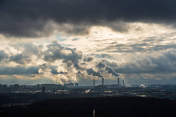 Wide view of an urban industrial area with several chimneys releasing smoke into a turbulent cloudy sky, with dark storm clouds above and bright gaps of light on the horizon, illustrating air pollution and environmental impact. Suitable for topics of climate change, industrial emissions, fossil fuel energy and ecology, with strong mood and ample horizontal copy space in the sky.