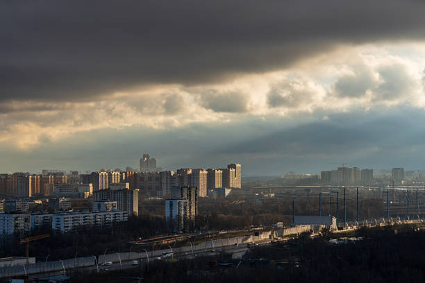 Wide view of a residential city district with rows of apartment buildings and roads in the foreground, lit by dramatic sun rays shining through heavy dark storm clouds. Suitable for themes of urban landscape, weather, dramatic sky and city infrastructure, with horizontal orientation and ample copy space in the cloudy sky.