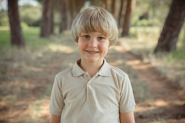 Smiling blond boy standing outdoors in a forest, natural light portrait, happy child looking at the camera.