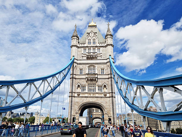 London, United Kingdom - 2024-08-20:  Crowds of pedestrians and vehicles cross the iconic Tower Bridge on a bright summer day, with the bridge's central stone tower and blue suspension cables framed against a partly cloudy sky in the historic heart of the city.